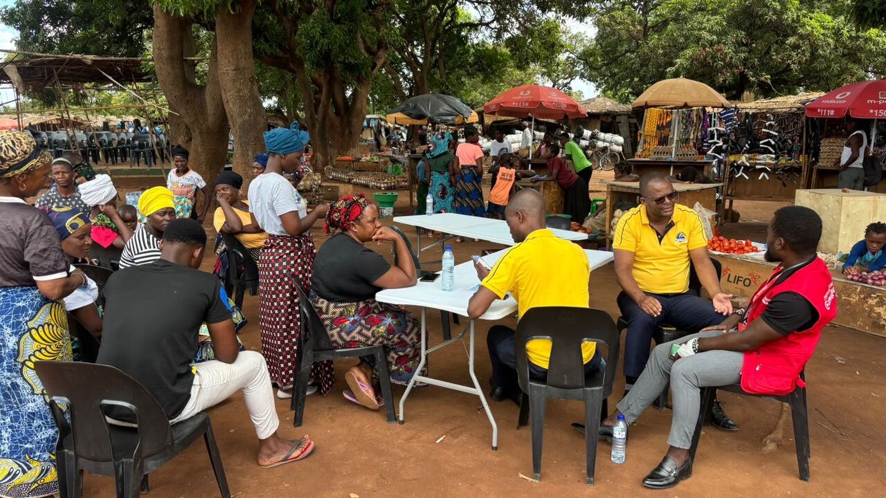 Reunião ao ar livre entre pessoas de camisola amarela e vendedoras, junto às bancas do mercado.